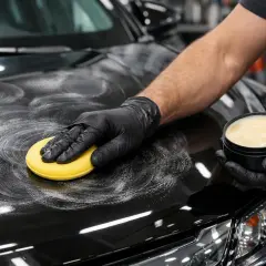 Ręka w czarnej rękawiczce nakłada wosk na czarną maskę samochodu. Czy po woskowaniu płukać auto? To pytanie zadaje sobie wielu kierowców.