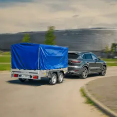 Szary SUV z przyczepą pod niebieską plandeką jedzie z dużą prędkością, mijając nowoczesny budynek.