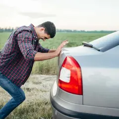 Mężczyzna w kratę próbuje odpalenie na pych zepsutego samochodu na tle pola.