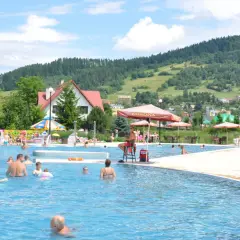 Słoneczny dzień w **Tylicz baseny termalne**. Ludzie kąpią się i relaksują w basenie, w tle góry i domy.