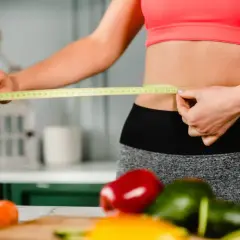 Mujer mide su cintura con cinta métrica, rodeada de verduras frescas. ¿La menopausia adelgaza o engorda?
