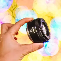 Dłoń trzyma obiektyw Canon 50mm na tle kolorowych świateł bokeh.