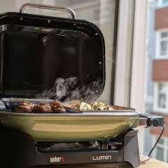 Leckeres Grillen auf dem Balkon: Gefüllte Champignons und Spieße garen in einem grünen Weber Lumin Grill. Rauch steigt auf.