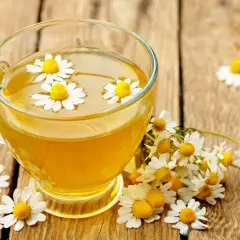 Une tasse de tisane dorée avec des fleurs de camomille flottantes, entourée de fleurs fraîches. Un rappel des bienfaits apaisants de la camomille.