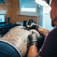 Artysta tworzy skomplikowany tatuaż na plecach klienta. Widać detale wzoru i ręce tatuażysty w czarnych rękawiczkach.