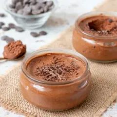 Deux pots de mousse au chocolat, l'un garni de copeaux de chocolat noir, sur une toile de jute. Une cuillère dorée présente une portion de cette délicieuse mousse.