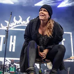 Beth Hart na scenie, w czapce i swetrze, śpiewa swoje utwory. W tle logo z koroną i perkusista.