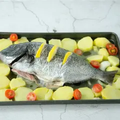 Un sargo pez entero, listo para hornear, rodeado de rodajas de patata y tomates cherry en una bandeja.