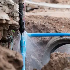 Wyciek wody z niebieskiej rury, pokazujący problematyczne przyłącze wody krok po kroku. Woda tryska na ziemię i betonowe rury.