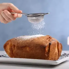 Una mano setaccia zucchero a velo su un dolce appena sfornato, un tocco finale perfetto per ogni ricetta Bimby.