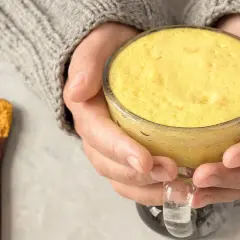 Warme goldene Milch in einer Tasse, gehalten von Händen in einem Strickpullover. Daneben ein Löffel mit Kurkumapulver. Die goldene Milch Wirkung ist bekannt für ihre entzündungshemmenden Eigenschaften.