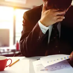 Pracownik w stresie, przykład sytuacji kryzysowych. Zmęczony mężczyzna w garniturze z ręką na twarzy, przy biurku z dokumentami i kubkiem kawy.