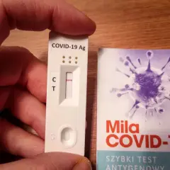 Ręka trzyma test antygenowy COVID-19 z jedną kreską. Obok opakowanie z grafiką wirusa i napisem "Szybki test antygenowy".