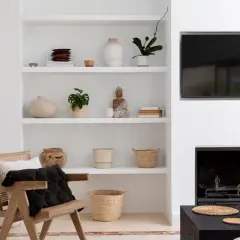 Elegancka aranżacja półki w salonie z ceramiką, roślinami i figurką Buddy. Obok wygodny fotel i kominek.