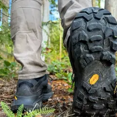 Close-up of a person's hiking boots on a forest trail, showcasing the aggressive tread pattern and the iconic yellow Vibram sole logo, highlighting the durability and grip of various Vibram sole types.