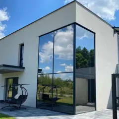 Nowoczesny dom z imponującym, narożnym oknem weneckim, w którym odbija się błękitne niebo z chmurami i zieleń drzew.