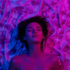 Donna con occhi chiusi, immersa in un letto di seta e petali, illuminata da luci blu e rosa. Un'immagine che evoca il piacere e il mistero del perché godiamo quando veniamo.