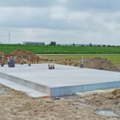 Gotowy projekt płyty fundamentowej na budowie. Betonowa płyta z wystającymi rurami i zbrojeniem, otoczona piaskiem i ziemią.