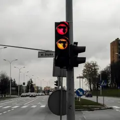 Sygnalizator S3 z czerwonym i żółtym światłem kierunkowym w lewo, na tle pochmurnego nieba i bloków mieszkalnych.