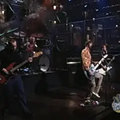 Zespół gra na scenie, muzycy chcą nauczyć się latać, a basista w czerni i gitarzyści w kolorowych koszulach tworzą energię.