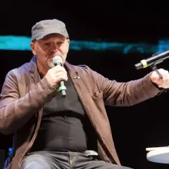 Uomo con cappellino e giacca di pelle marrone parla al microfono, sembra che sono ancora in coma, ma è sul palco.