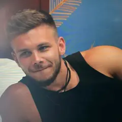Bartek z Hotel Paradise, z uśmiechem na twarzy, w czarnej koszulce, na tle tropikalnej tapety.