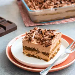 Una fetta di tiramisù cioccolato, ricca e cremosa, guarnita con riccioli di cioccolato. Accanto, pezzi di cioccolato fondente e un'intera teglia.