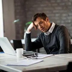 Zmęczony mężczyzna przy biurku zastanawia się, skąd wziąć pieniądze na już. Laptop, dokumenty i kubek kawy sugerują trudny dzień.