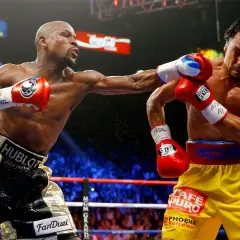 Two boxers in a ring, one throwing a punch. The inside fighter, with gold shorts, connects with his opponent.