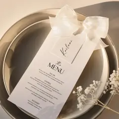 Menu de mariage élégant sur une assiette, orné d'un ruban et de petites fleurs blanches.