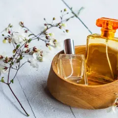 Dwie buteleczki perfum, jedna w szkle, druga w bursztynowym kolorze, w drewnianej miseczce, otoczone delikatnymi kwiatami. Czy to woda toaletowa czy tester?