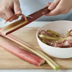 Hände schälen Rhabarberstangen auf einem Holzbrett. Die abgezogenen Streifen landen in einer Schüssel.