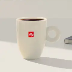 Tazza bianca con logo Illy, piena di caffè americano, pronta per una pausa studio a casa.