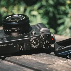 A Fujifilm X-Pro3 camera with a lens and lens hood rests on a wooden surface, ready for a photography review.