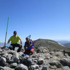 Dos excursionistas celebran en la cima tras una **cuerda larga ruta**. Un poste verde marca el camino.