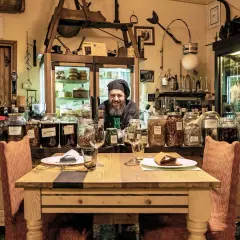 Un cuoco sorridente in un locale con salumi appesi e barattoli di conserve. Un'atmosfera unica per ristoranti particolari a Brescia e provincia.