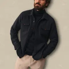 Un homme porte une surchemise foncée ouverte sur un col roulé bleu marine, avec un pantalon beige. Idéal pour savoir comment porter une surchemise avec style.