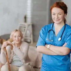 Uśmiechnięta pielęgniarka w niebieskim uniformie, zafascynowana kursem opiekuna medycznego, stoi obok starszej pani z laską.