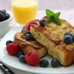 Tostadas francesas doradas, decoradas con arándanos y frambuesas frescas, junto a un vaso de jugo de naranja y un tazón de bayas.