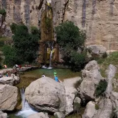 Un hermoso salto de agua cae en una poza natural rodeada de rocas y vegetación. Varias personas disfrutan del paisaje y del agua.