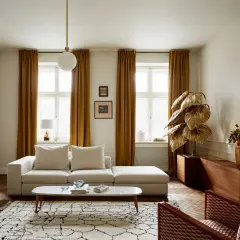 Przytulny salon w kamienicy z beżową kanapą, złotymi zasłonami i ozdobną rośliną.