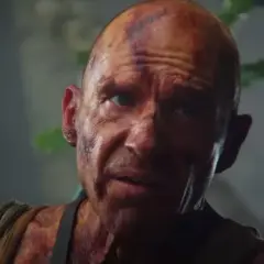 Ralph Fiennes, med smuts och skador i ansiktet, talar intensivt med en ung person i en grönskande, dimmig miljö.