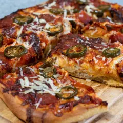 En saftig pan pizza med pepperoni och jalapeños, toppad med riven ost, ligger på en träskärbräda.