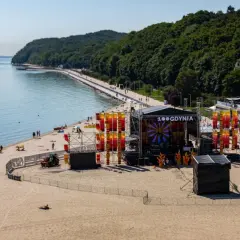 Scena festiwalowa na plaży w Gdyni, z widokiem na morze i las. Kolorowe dekoracje i logo "100 Gdynia" na scenie.
