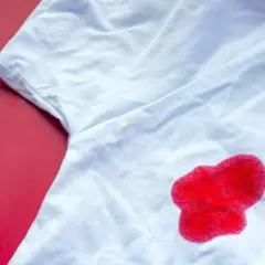 Biała koszulka z plamą z czerwonego płynu i kilkoma mniejszymi, żółtymi plamkami.