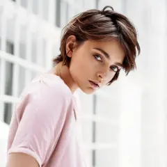 Junge Frau mit einem lässigen Pixie-Cut-Haarschnitt, blickt nachdenklich in die Kamera. Sie trägt ein zartrosa Oberteil.