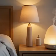 Un difusor emite vapor, ayudando a como deshumidificar una habitación. Junto a una lámpara y un frasco en una mesita de noche.