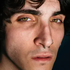 Primo piano di un giovane con occhi verdi penetranti, capelli scuri e piercing al naso. Giuse the Lizia, un volto intenso.
