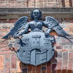 Kamienna rzeźba anioła z kluczem i tarczą z zamkiem, symbolizująca herb Torunia, zdobi ceglaną ścianę.
