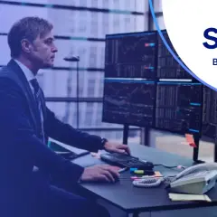 Mężczyzna w garniturze analizuje wykresy na monitorach w biurze Saxo Bank Polska. "Be invested" - hasło zachęca do inwestowania.